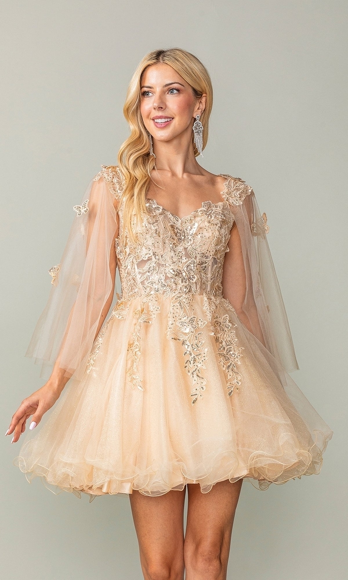 A woman stands against a plain background wearing the Butterfly-Cape Short A-Line Homecoming Dress 3393, featuring light gold tulle, sheer flared sleeves, and delicate sequin appliqués.